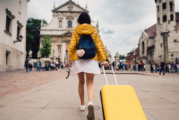 Turista de costas com mala amarela caminhando sobre um cenário que remete o Centro de uma cidade.